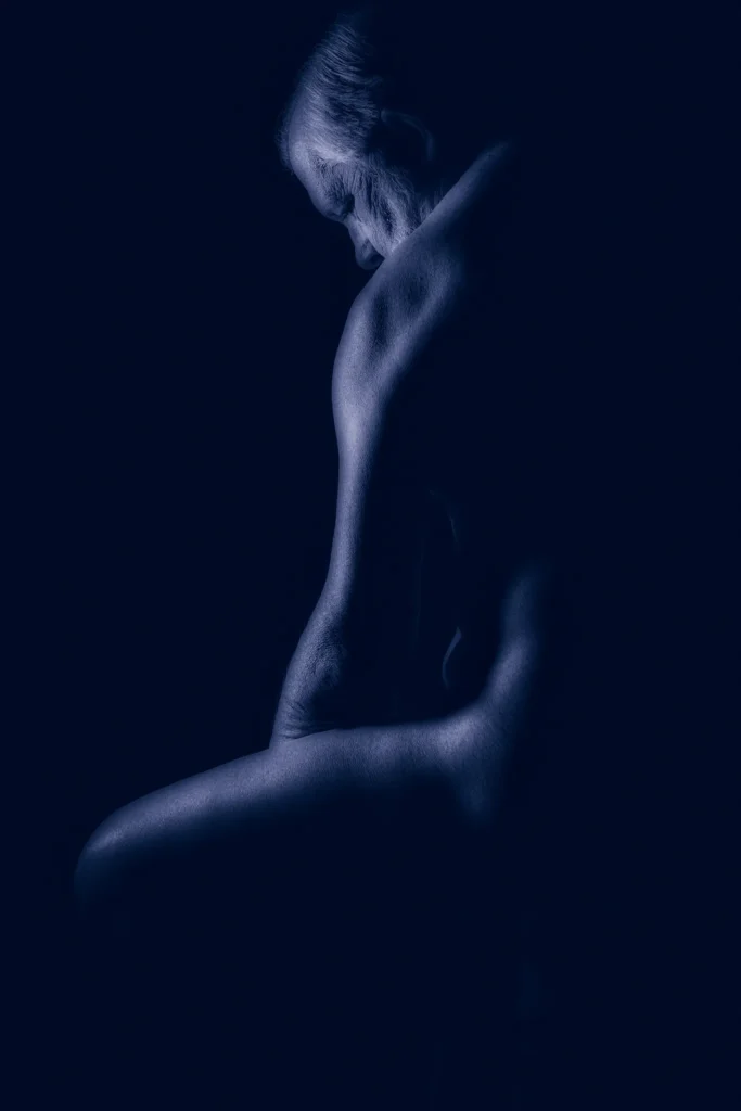 Blue-toned fine art image of Larry seated in profile with head lowered, defined by soft directional light against a dark background.