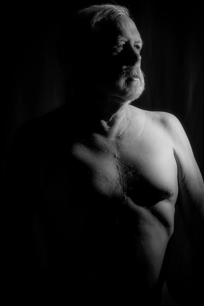 Black and white portrait of Larry with bare torso, looking upward in dramatic side lighting against a dark background.