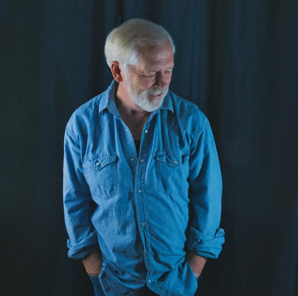 Lifestyle portrait of Larry wearing a blue denim shirt, standing with hands in pockets and looking down against a dark curtain background.