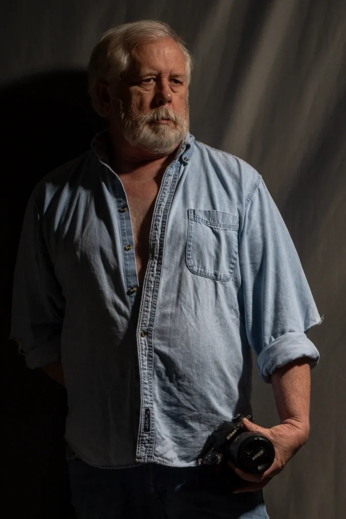 Lifestyle portrait of Larry wearing a light denim shirt and holding a Nikon camera in dramatic side lighting.