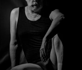 Seated Figure in Shadow Black and white fine art image of a seated mature male model in dramatic side lighting against a dark background.