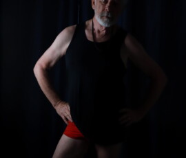 Studio Pose in Black and Red
Photo by Rebel Jones Portrait of Larry wearing a black tank top and red briefs, standing with hands on hips against a dark curtain background.