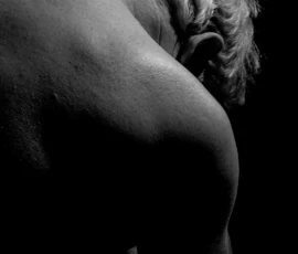 Back Study in Shadow Black and white bodyscape of a male model’s back and shoulder with head lowered, emphasizing curved form, skin texture, and deep shadow.