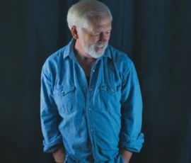 Denim Portrait
Photo by Rebel Jones Lifestyle portrait of Larry wearing a blue denim shirt, standing with hands in pockets and looking down against a dark curtain background.