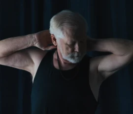 Quiet Strength
Photo by Rebel Jones Portrait of Larry wearing a black tank top with arms raised behind his head, looking downward against a dark curtain background.